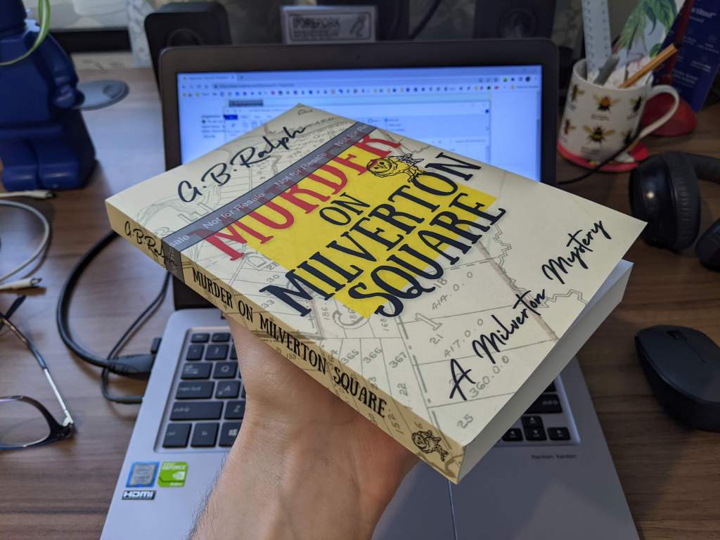 Murder on Milverton Square (A Milverton Mystery) by G B Ralph. Paperback proof held up in front of a laptop.