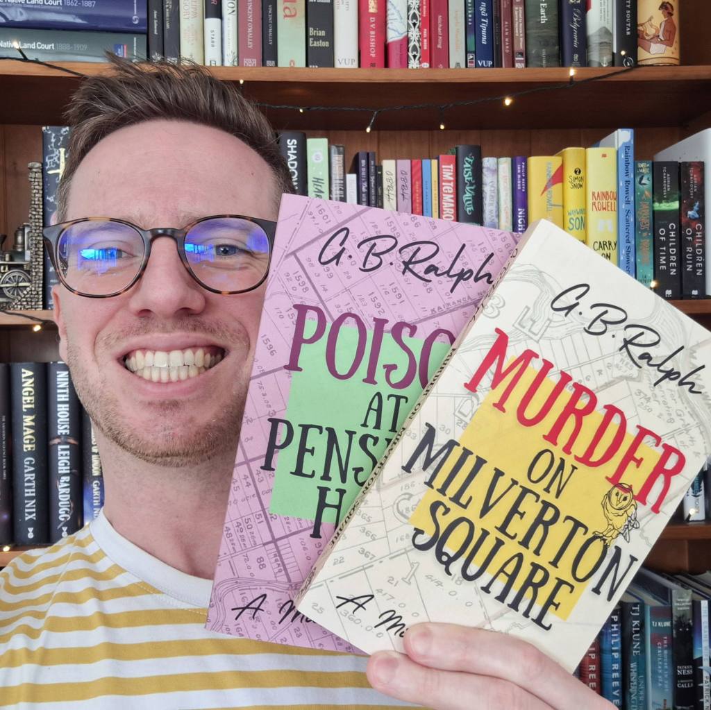 Author G. B. Ralph holding up paperback copies of Murder on Milverton Square and Poison at Penshaw Hall