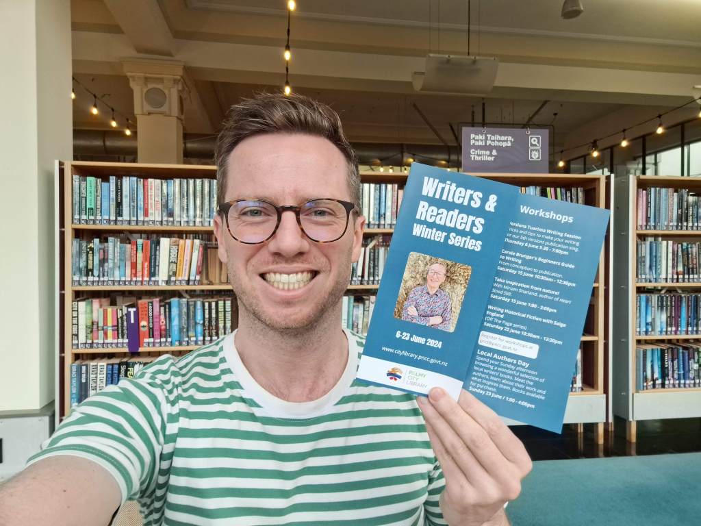 Author G B Ralph in front of book shelves at the Palmerston North City Library, holding a pamphlet titled "Writers and Readers Winter Series"