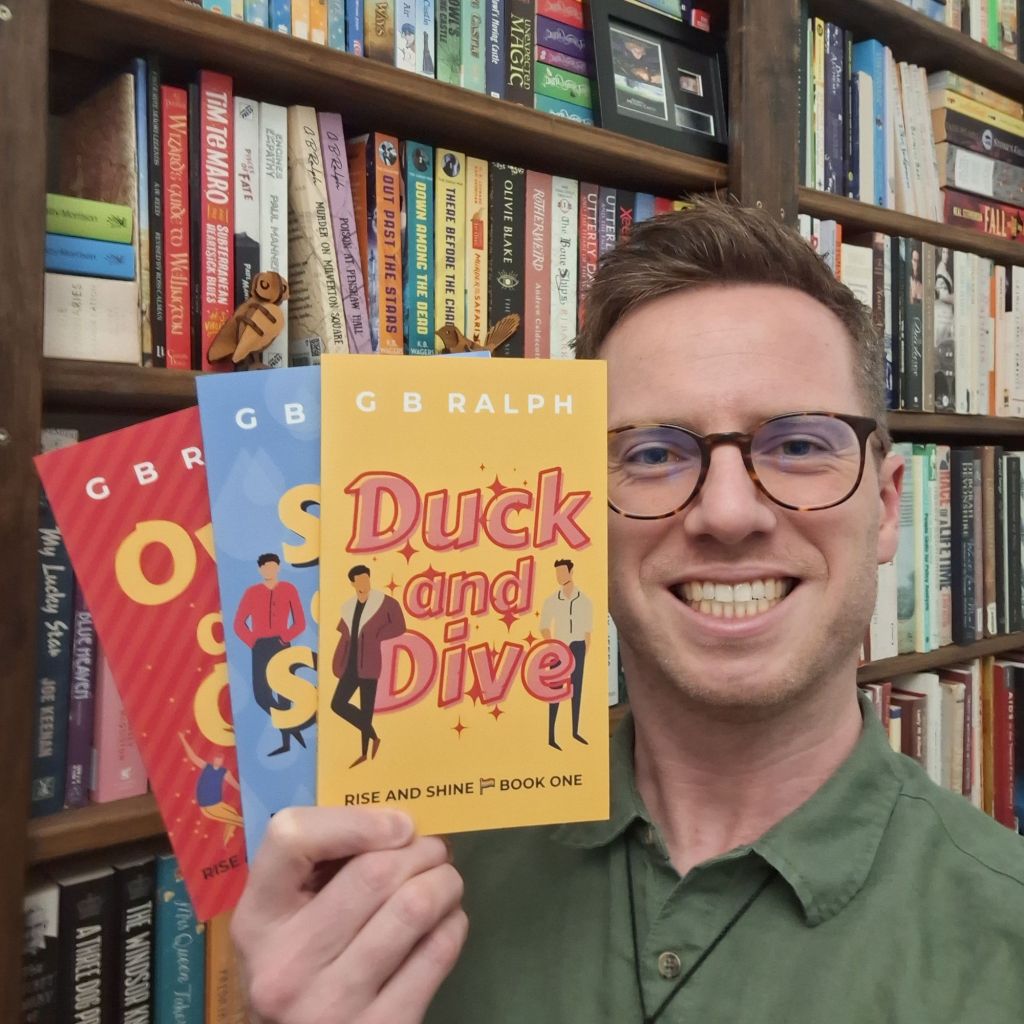 Author holding up books in front of bookshelves. First book is Duck and Dive by G B Ralph.