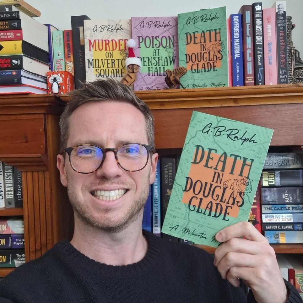 Selfie with book in hand and bookshelf in background. Death in Douglas Glade by G B Ralph.