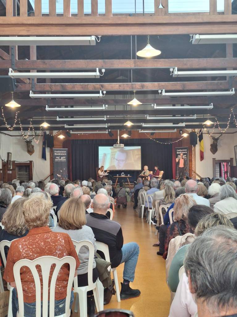 Hall full of people seated and listening to a panel on stage including moderator, two authors in person, one author on big screen on video call.