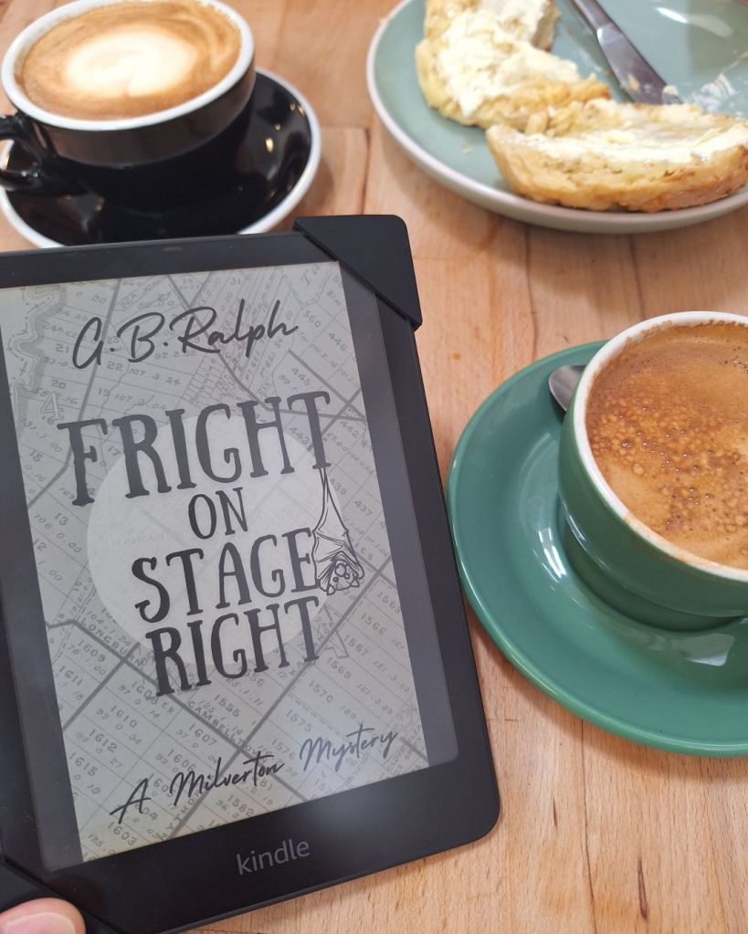 Kindle ereader displaying book cover of Fright on Stage Right by G. B. Ralph. In the background: a cafe table with coffees and scone.
