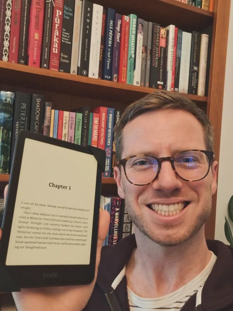 Selfie in front of a bookcase while holding an ereader displaying Chapter 1