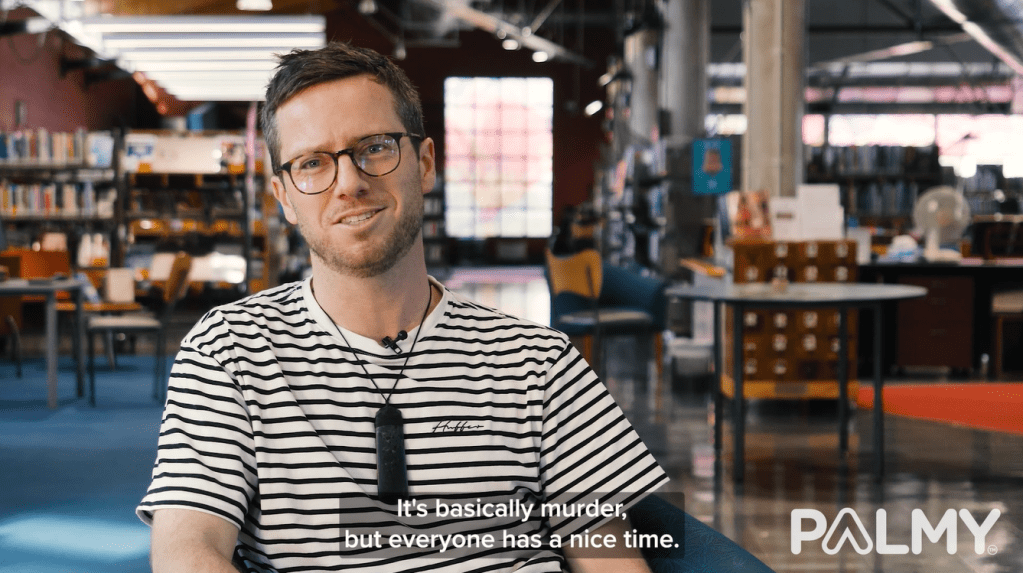 Video screenshot of author G. B. Ralph sitting and speaking to the camera with the library in the background. Subtitle reads "It's basically murder, but everyone has a nice time."