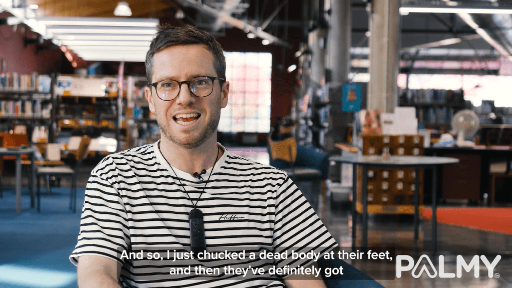 Video screenshot of author G. B. Ralph sitting and speaking to the camera with the library in the background. Subtitle reads "And so, I just chucked a dead body at their feet and then they've definitely got—"