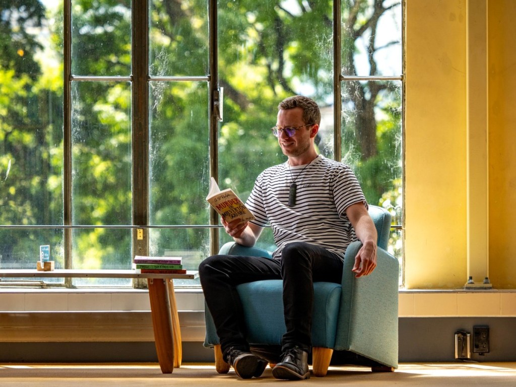 Author G. B. Ralph pretending to read Murder on Milverton Square while sitting in front of windows.